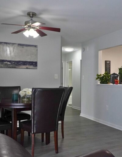 Interior of a modern dining room with a dark wood table, four chairs, a ceiling fan above, and decorative plants visible through a window pass-through to the kitchen.